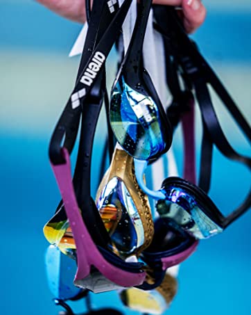 athlete holding several pairs of arena goggles, in blue, pink, gold and black, above a pool